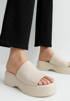 Person iført sorte bukser og beige platform-sandaler står indendørs på et lyst gulv. Kun underben og fødder er synlige.