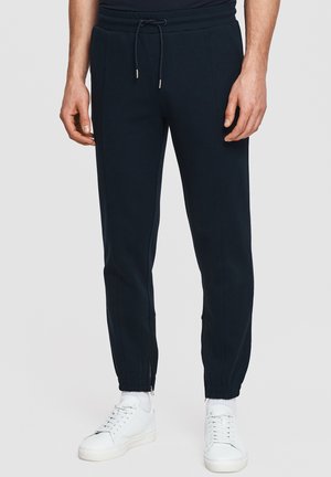 Mężczyzna noszący czarne spodnie dresowe ze ściągaczem w pasie i suwakami przy kostkach, w zestawieniu z białymi trampkami i skarpetkami.