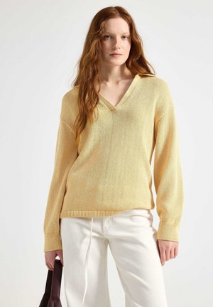 Lys gul strikket sweater med V-udskæring, lange ærmer og en afslappet pasform. Ribbet mansjetter og kant; parret med hvide bukser og en brun taske.