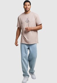 Camiseta de algodón rosa claro con cuello redondo, que presenta el texto "inocente hasta que se demuestre lo contrario" en gris oscuro, combinada con pantalones vaqueros de denim azul claro.