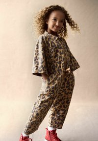 Beige ja brown leopardikuvioinen jumpsuit, lyhyet hihat, vetskarikiinnitys, etutaskut, yhdistetty punaisiin korkeakorkoisiin tennareihin ja valkoisiin sukkiin.