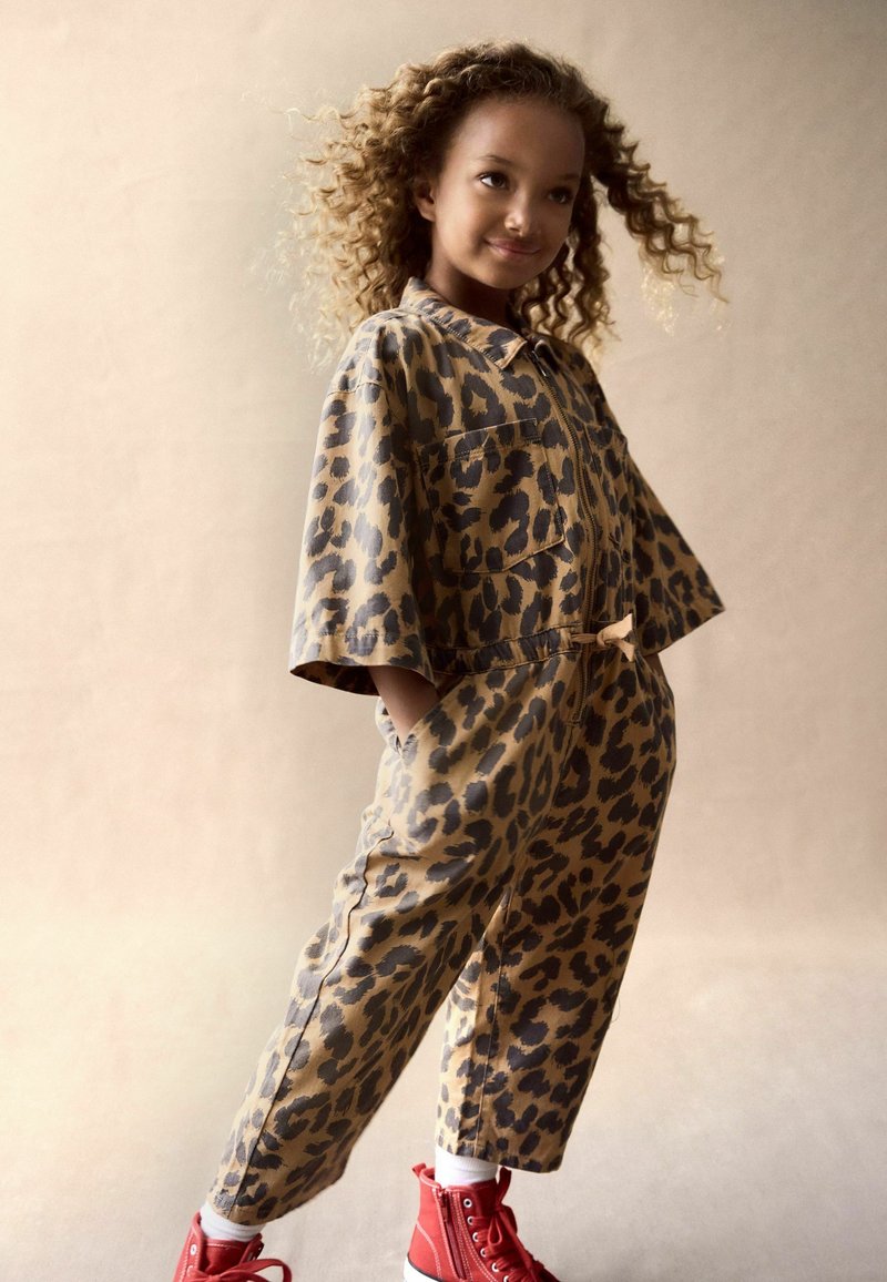Beige ja brown leopardikuvioinen jumpsuit, lyhyet hihat, vetskarikiinnitys, etutaskut, yhdistetty punaisiin korkeakorkoisiin tennareihin ja valkoisiin sukkiin.