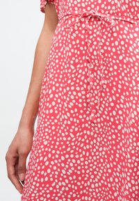 Κοραλί φόρεμα με λευκές βουλίτσες, κοντά μανίκια, ζώνη στη μέση και μαλακή υφή υφάσματος. Διαθέτει άνετη εφαρμογή και διακριτικό βολάν στο τελείωμα.