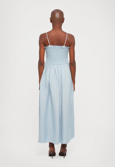 Lichtblauwe maxidress met een gepast, geschroefd lijfje en een gerimpelde halslijn. Mouwloos ontwerp dat overgaat in een losse rok. Geklede schoenen.