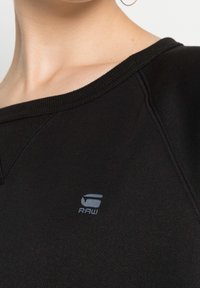 Felpa nera con scollatura ampia, caratterizzata da dettagli cuciti e un piccolo logo grigio "RAW" sul petto. Tessuto morbido e con una texture.