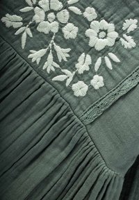 Grünes Kleidungsstück mit floralem weißen Stickmuster, das über eine gepufferte Textur und Spitzen-Details verfügt. Weicher, leichter Stoff mit dezentem Glanz.