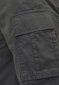 Tmavě šedý cargo jacket rukáv se velkou kapsou s klopou, vyztuženým prošíváním a hladkým, matným povrchem.