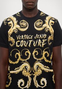 Black T-shirt featuring intricate gold swirling patterns and bold text "VERSACE JEANS COUTURE" in white, made from soft fabric.