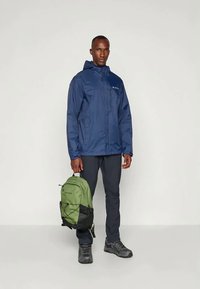 Marineblauer wasserdichter Jacke mit Kapuze, kombiniert mit schwarzen Hosen und einem grünen Rucksack. Verfügt über einen Frontreißverschluss und ein sichtbares Markenlogo.