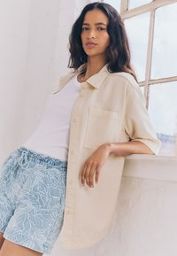 Camicia beige chiaro con bottoni e tasca, indossata sopra una canottiera bianca e pantaloncini blu con motivi di foglie ricamate bianche.