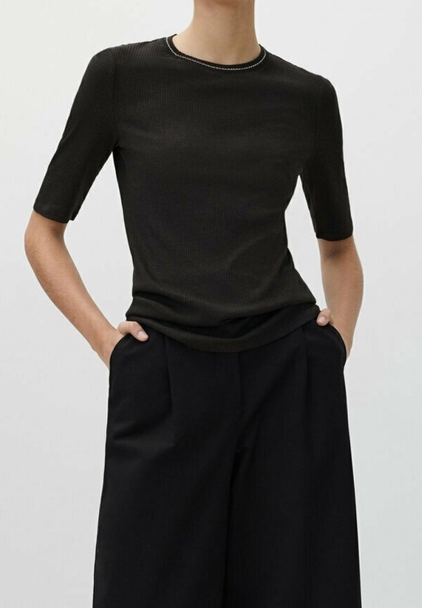 Personne portant un t-shirt noir côtelé ajusté à manches courtes et un pantalon noir ample taille haute, les mains dans les poches, fond neutre.