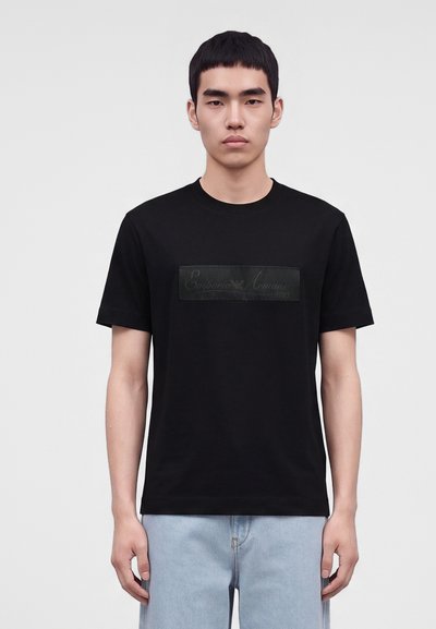 Млад мъж с къса черна коса, облечен с черна тениска Emporio Armani и светлосини дънки на едноцветен сив фон.