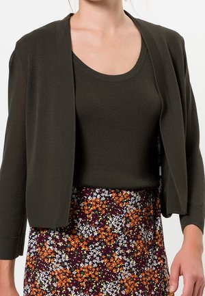 Femme portant un cardigan vert foncé et un haut associés à une jupe taille haute avec un petit imprimé floral orange, blanc et violet.