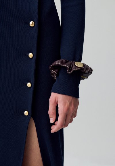 Abito a coste blu navy con bottoni dorati e una fessura alta fino alla coscia; presenta un gomitolo di pelle marrone con un accento dorato sul polso.