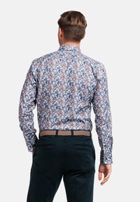 Chemise à manches longues florale avec un motif bleu, blanc et marron ; col structuré ; coupe ajustée ; associée à un pantalon foncé et une ceinture tissée.