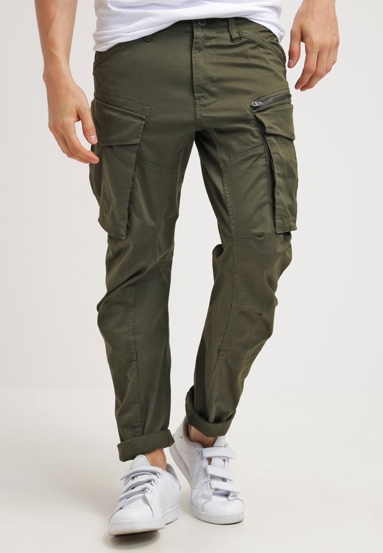Homme portant un pantalon cargo vert olive avec plusieurs poches, ourlets roulés, baskets blanches et une chemise blanche, debout devant un fond uni.