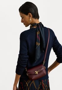 Žena oblečená v námorníckom pletenom svetri a potlačenej čiernej šatke previazanou okolo krku, nesúca tmavočervenú koženú cez telo tašku so zlatým uzáverom.