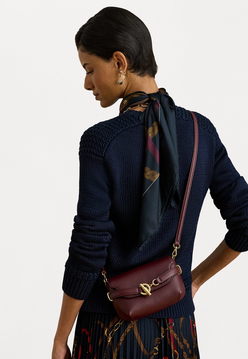 Žena oblečená v námorníckom pletenom svetri a potlačenej čiernej šatke previazanou okolo krku, nesúca tmavočervenú koženú cez telo tašku so zlatým uzáverom.