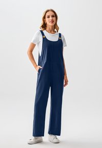 Marineblaue Overalls mit strukturierter Oberfläche, weitem Bein, Seitentaschen und Holzknöpfen an den Trägern, kombiniert mit einem weißen Hemd und weißen Turnschuhen.