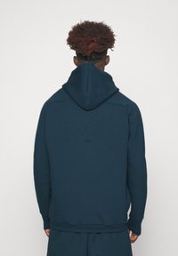 Person with short curly hair facing away, wearing a dark blue hooded sweatshirt and matching pants against a plain background.