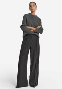 Grauer Strickpullover mit geripptem Muster und überschnittenen Schultern, kombiniert mit hoher Taille und weitem Bein in dunkelgrauer Hose. Glatte Lederschuhe.
