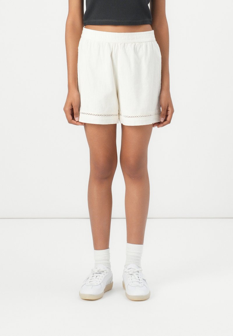 White cotton shorts with an elastic waistband and decorative stitching at the hem. Paired with white sneakers and ribbed socks.
