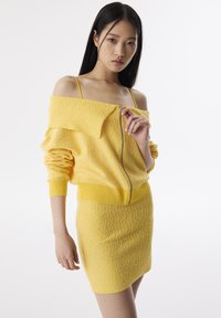 Ensemble coordonné tricoté jaune comprenant un haut zippé décolleté en tissu texturé et une mini-jupe ajustée, les deux ornés d'accents côtelés.