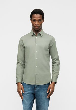 Moški v svetlo zeleni srajci z dolgimi rokavi in modrih kavbojkah stoji pred preprostim svetlim ozadjem.