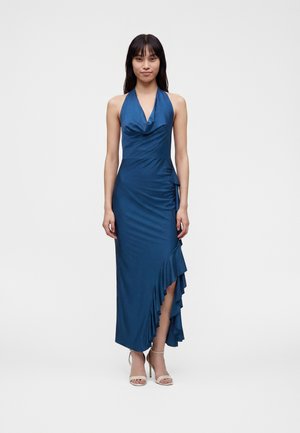 Femme portant une robe bleue sans manches avec un détail à volants et une fente haute, debout devant un fond blanc uni.