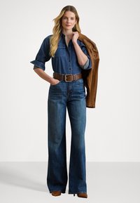 Denimskjorte med oppbrettede ermer, mørk vask, kombinert med high-waisted vide jeans og en brun lærjakke slengt over den ene skulderen.
