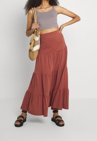 Jupe maxi tiered marron en tissu doux, associée à un top tank gris cropped et un sac en osier. Des sandales noires à lanières complètent le look.