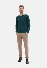 Pull à col en V bleu canard, pantalon beige et chaussures noires. Le pull a des poignets côtelés ; le pantalon est droit avec les manches retroussées. Design minimal.