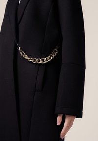 Il cappotto di lana nero presenta un design strutturato, una vestibilità over-size e una notevole cintura in catena dorata che mette in risalto la vita. Texture liscia in tutto il capo.