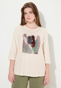 Helle beige T-Shirt mit Rundhalsausschnitt, versehen mit einem bunten gestickten Design von Bäumen in Grün, Kastanienbraun und Pink, mit einer weichen Textur.