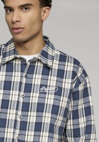 Jeune homme portant une chemise à carreaux bleue et blanche avec un logo brodé sur la poitrine gauche.