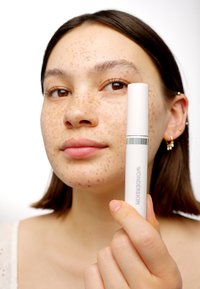 Tubetto bianco di prodotto Wonderskin tenuto davanti al volto di una persona, caratterizzato da lentiggini sparse e un tono di pelle neutro. Design semplice e cilindrico.