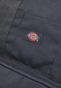 Tissu noir en gros plan montrant une étiquette carrée cousue avec le logo coloré Dickies et des détails de sangle solides.