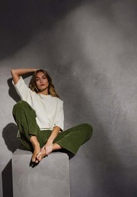T-shirt blanche surdimensionnée et pantalon large en velours côtelé vert, assis sur un piédestal en béton, avec un éclairage doux et un fond texturé.