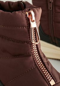 Stivali isolati marroni con una texture trapuntata, dotati di una zip rosa oro prominente e una suola nera a contrasto.