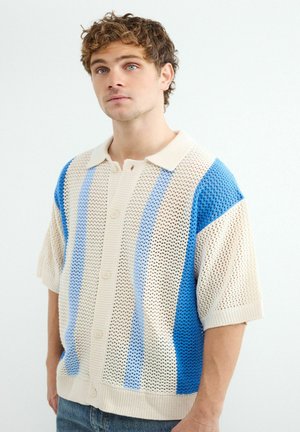 Młody mężczyzna z kręconymi włosami, ubrany w krótki, otwarty kardigan z pionowymi niebieskimi i kremowymi paskami.