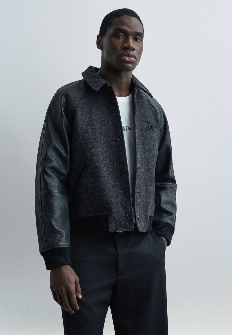 Άνδρας που φοράει σκούρο γκρι σακάκι με μαύρα δε ρμάτινα μανίκια, λευκό μπλουζάκι με γραφικό σχέδιο και μαύρο παντελόνι, στέκεται μπροστά σε ένα απλό ανοιχτό φόντο.