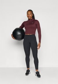 Burgunderfarget langermet crop topp med glidelås, matchende svarte høyspent leggings. Modellen holder en svart medisinball og har på seg svarte joggesko.