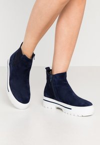 Námořnické modré nubukové kotníkové boty s bílou gumovou podrážkou. Obsahují zipové zapínání a texturovaný povrch. Elegantní design s minimálními akcenty.