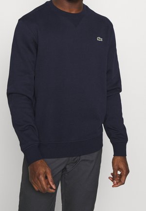 Man draagt een marineblauwe trui met ronde hals en lange mouwen, voorzien van een klein groen logo op de linkerborst, gecombineerd met donkergrijze broek.