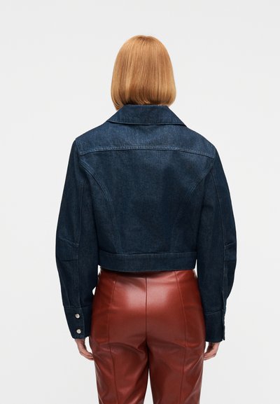 Άτομο με ίσια κόκκινα μαλλιά μέχρι τους ώμους φοράει ένα σκουρό μπλε τζιν μπουφάν και ψηλόμεσα καφέ δερμάτινα παντελόνια, φαίνεται από πίσω.