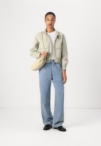 Veste cropped vert clair avec deux poches avant, t-shirt blanc en dessous, jean bleu ample et chaussures noires. Sac beige tenu à la main gauche.