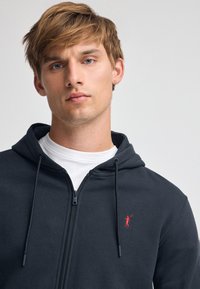 Granatowa bluza z kapturem na zamek błyskawiczny, z kieszenią z przodu, kapturem ze sznurkiem oraz małym czerwonym logo na lewej piersi, noszona na białej koszulce.