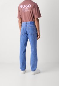 Jeans de denim de color azul claro con un ajuste relajado, cinco bolsillos y un parche con el logo en la parte trasera de la cinturilla. Llevado con una camiseta marrón de manga corta.