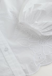 Valge puuvillane pluus kroogitud varrukatega, millel on keeruka lillemustriga tikand, tekstuuriga detailid ja väikesed nupud.