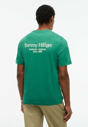 Mann trägt grünes Tommy Hilfiger T-Shirt mit weißem Text auf dem Rücken und beige Hose, schaut weg auf einfachem weißen Hintergrund.